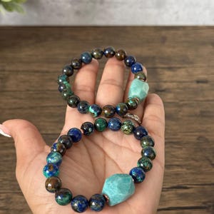 May include: Two bracelets made with blue, green, and brown gemstone beads. Each bracelet has a larger, light green gemstone bead in the center.