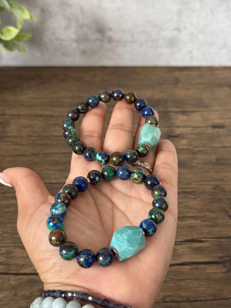 May include: Two bracelets made with blue, green, and brown gemstone beads. Each bracelet has a larger, light green gemstone bead in the center.