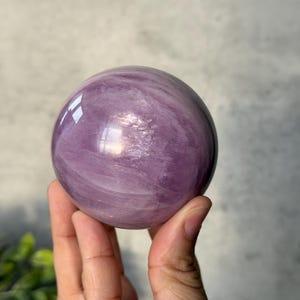 May include: A polished, spherical lavender-coloured crystal with visible internal veining and natural cloud-like inclusions. The surface of the stone is smooth and reflective, catching light to highlight the varying shades of purple throughout the orb.