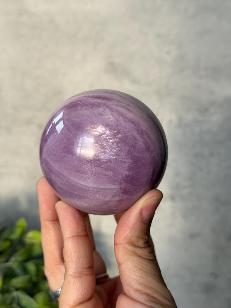 May include: A polished, spherical lavender-coloured crystal with visible internal veining and natural cloud-like inclusions. The surface of the stone is smooth and reflective, catching light to highlight the varying shades of purple throughout the orb.