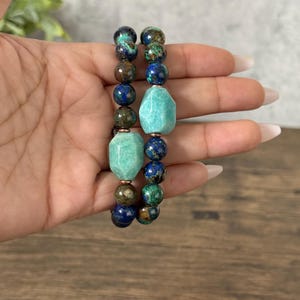 May include: Two bracelets made of blue, green, and brown gemstone beads. The bracelets feature larger, faceted, turquoise-colored beads.