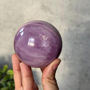 May include: A polished, spherical stone with a smooth, glossy surface. The object features a translucent, light purple colour with visible internal cloudy white veining and natural mineral inclusions throughout the rounded, orb-like structure.