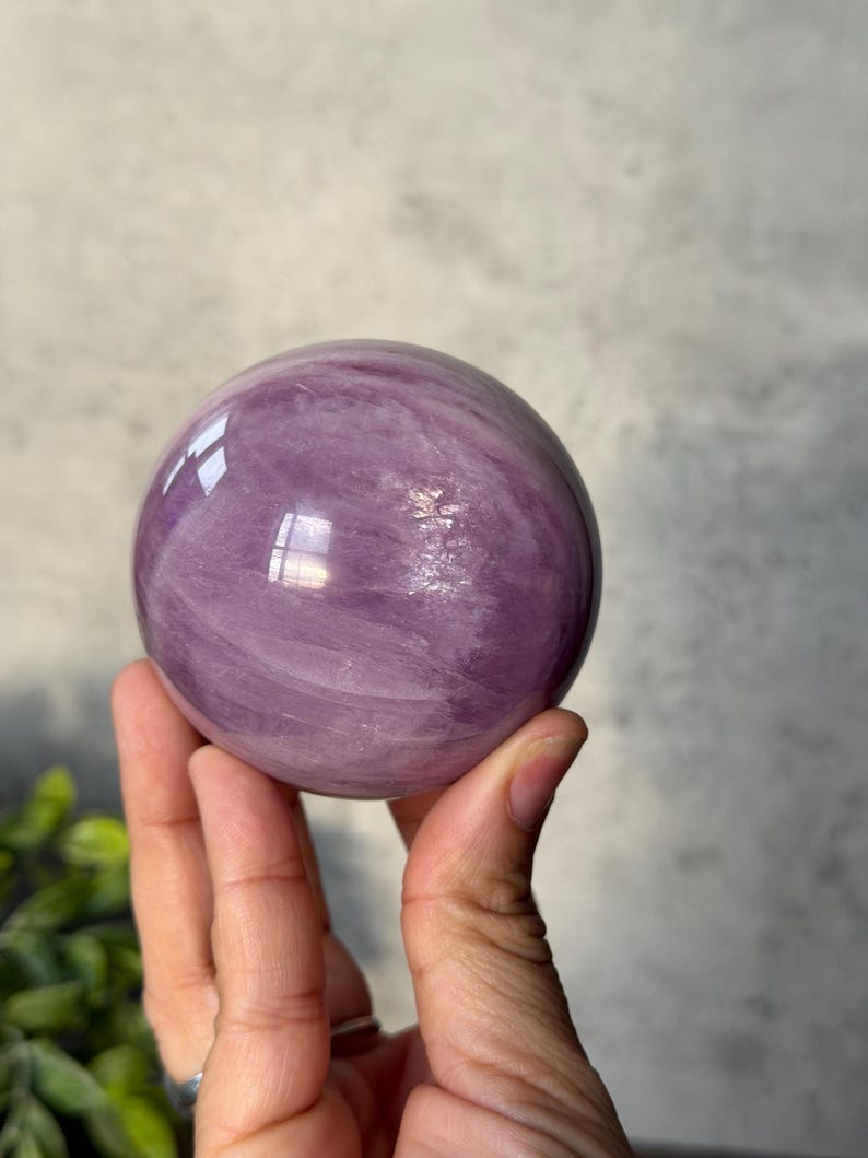 May include: A polished, spherical stone with a smooth, glossy surface. The object features a translucent, light purple colour with visible internal cloudy white veining and natural mineral inclusions throughout the rounded, orb-like structure.