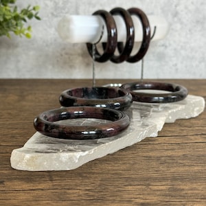 May include: Four dark red and black gemstone bracelets on a white crystal stand. The bracelets are stacked on top of each other and are arranged in a circle.