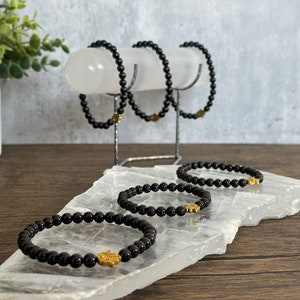 May include: Five black beaded bracelets with gold hamsa hand charms. The bracelets are displayed on a white crystal stand.