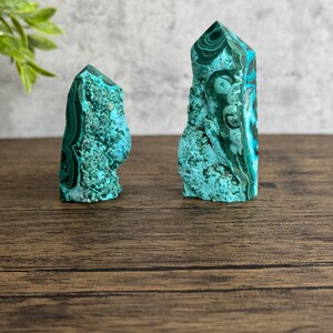 Puede incluir: Dos puntas de cristal de malaquita verde y azul sobre una superficie de madera. Los cristales son altos y delgados, con una punta puntiaguda y una superficie rugosa y texturizada.