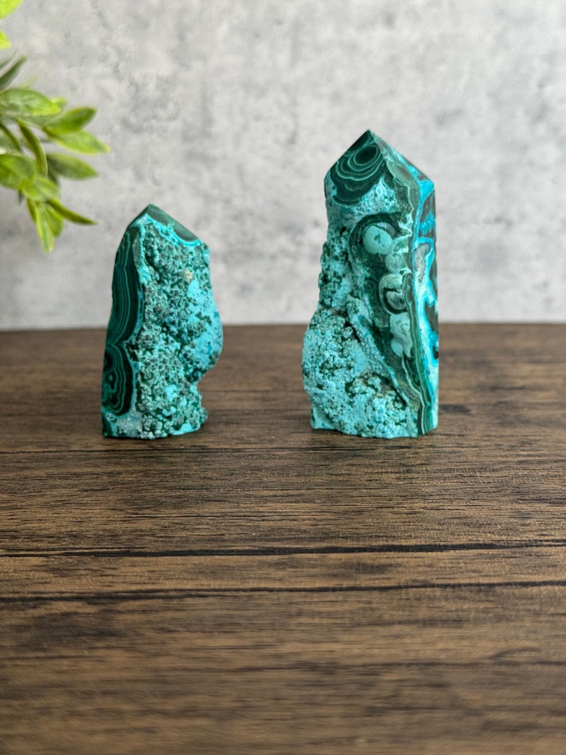 Puede incluir: Dos puntas de cristal de malaquita verde y azul sobre una superficie de madera. Los cristales son altos y delgados, con una punta puntiaguda y una superficie rugosa y texturizada.