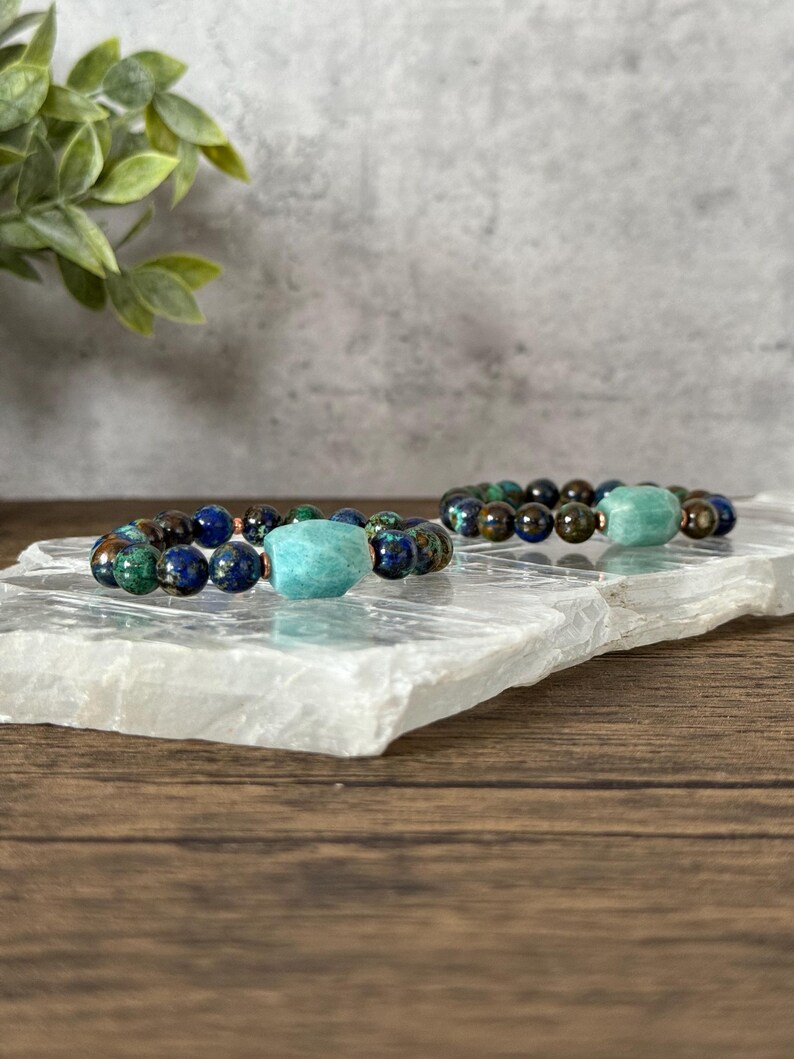 May include: Two beaded bracelets with a turquoise green stone accent. The bracelets are made with dark brown, blue, and green beads.