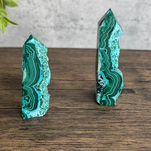 Puede incluir: Dos puntas de cristal de malaquita verde, una m&aacute;s grande que la otra, sobre una superficie de madera marr&oacute;n. Los cristales tienen un patr&oacute;n de bandas verdes y azules.