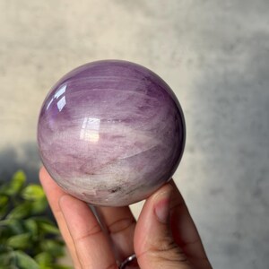 May include: A polished, spherical crystal ball featuring a smooth surface with swirling patterns of light purple, lavender, and white. The stone displays natural internal inclusions and reflective highlights across its rounded, glossy exterior.