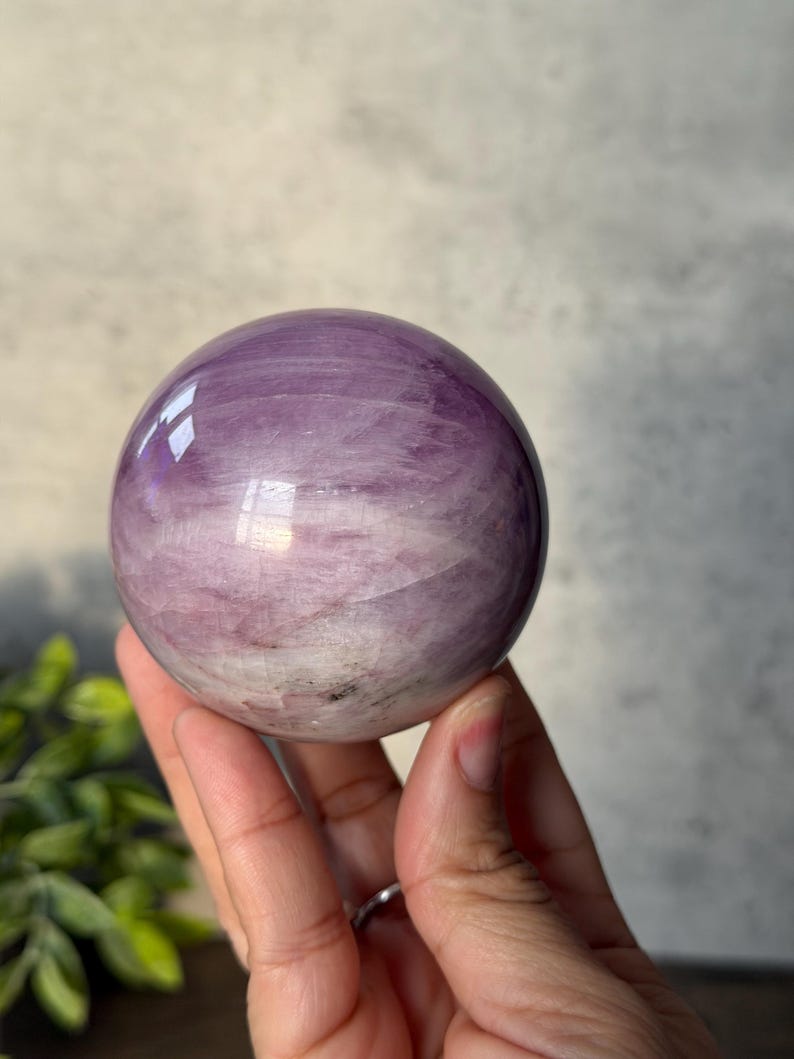 May include: A polished, spherical crystal ball featuring a smooth surface with swirling patterns of light purple, lavender, and white. The stone displays natural internal inclusions and reflective highlights across its rounded, glossy exterior.