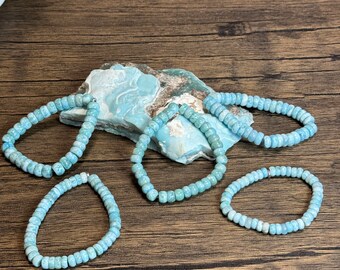 May include: Four turquoise blue gemstone bracelets made of small round beads. The bracelets are arranged on a wooden surface with a piece of blue and brown patterned stone in the center.