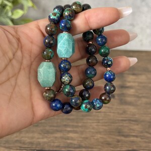May include: Three beaded bracelets with green, blue, and brown stones. The bracelets are stacked on top of each other on a hand.