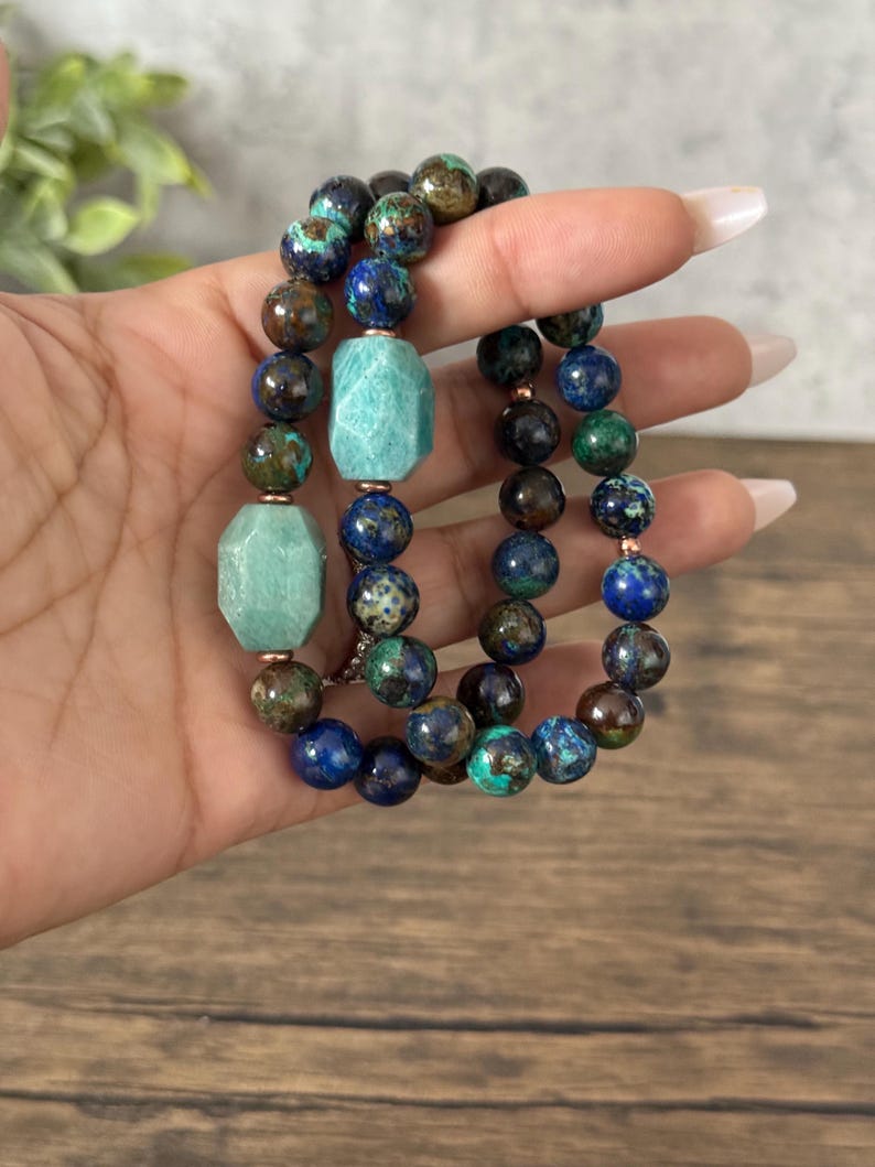 May include: Three beaded bracelets with green, blue, and brown stones. The bracelets are stacked on top of each other on a hand.