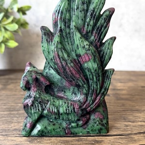May include: A carved zoisite figurine featuring a multi-tailed fox with prominent green and black speckled patterns and deep magenta inclusions. The sculpture displays a smooth, polished surface with intricate detailing on the fox's body and tails.