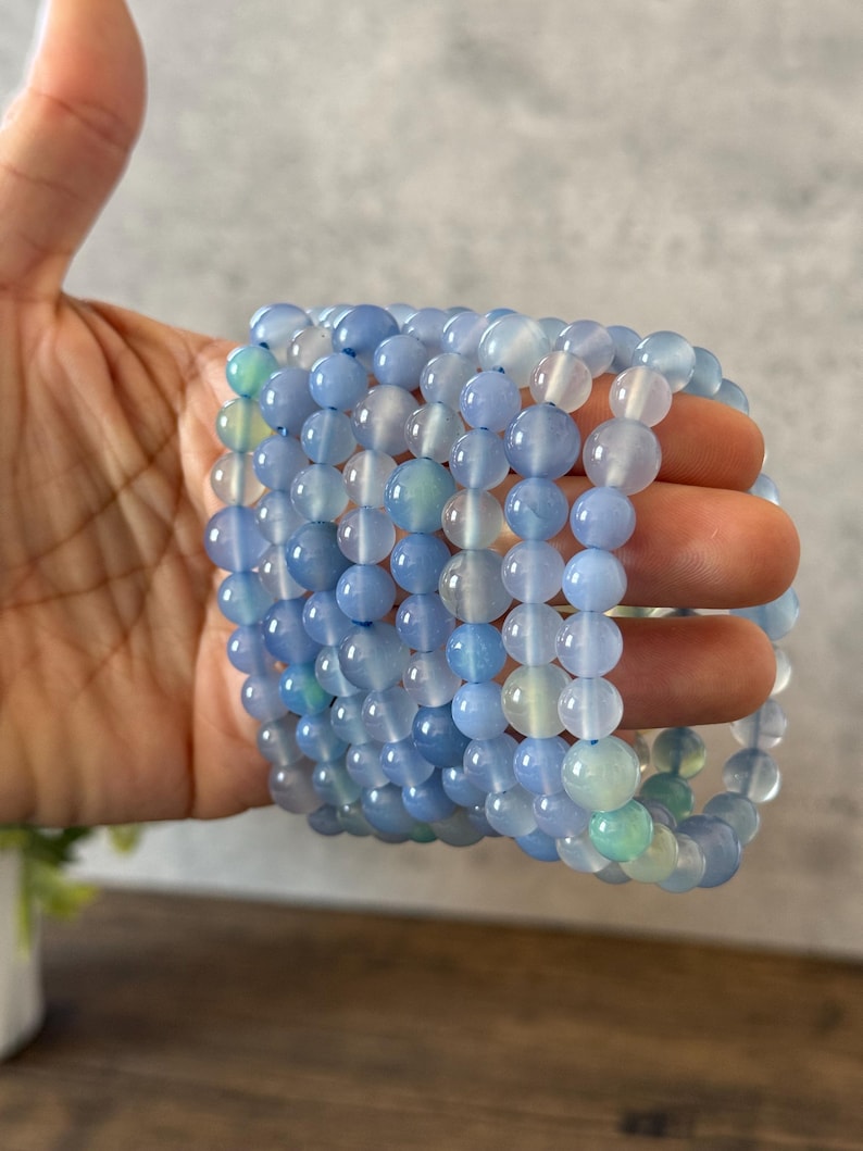 Puede incluir: Una pila de pulseras de &aacute;gata azul con un degradado de color azul claro y blanco. Las pulseras est&aacute;n hechas de cuentas redondas y est&aacute;n apiladas unas sobre otras.