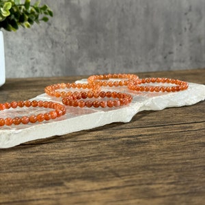 May include: Four orange carnelian gemstone bracelets stacked on a white selenite slab. The bracelets are made of small, round beads.