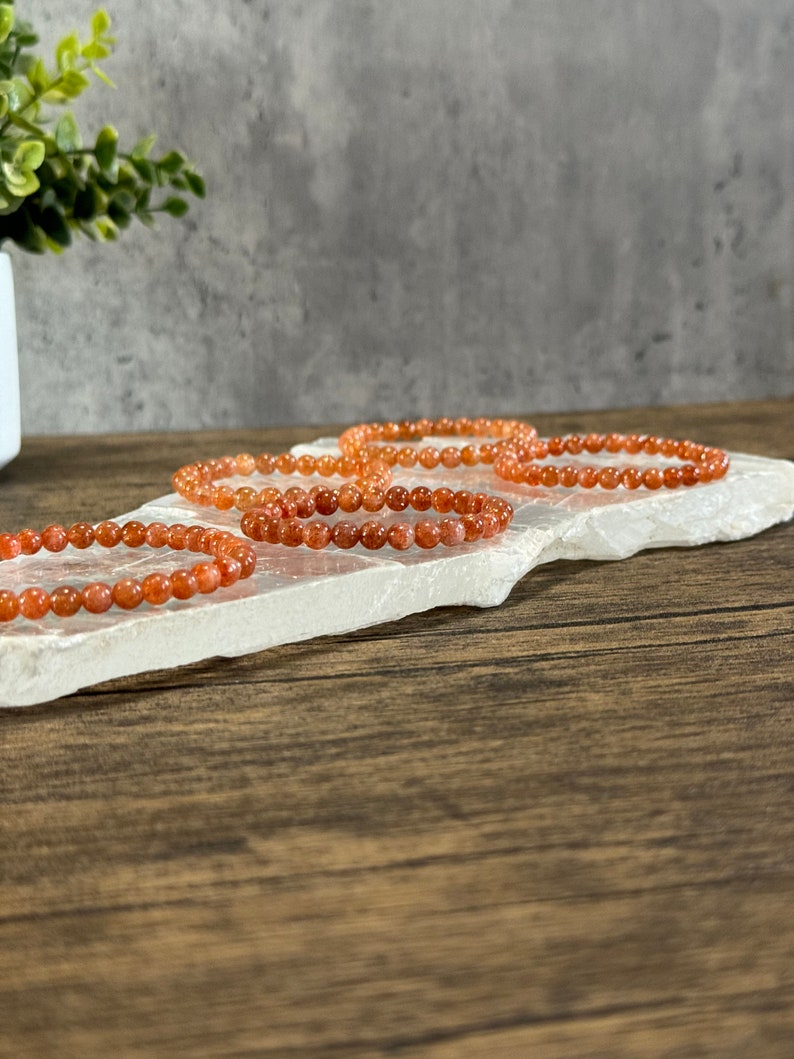 May include: Four orange carnelian gemstone bracelets stacked on a white selenite slab. The bracelets are made of small, round beads.