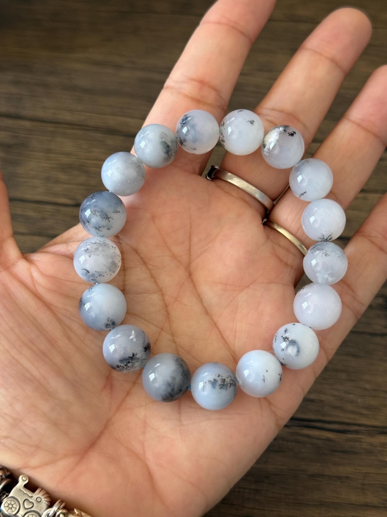 May include: A white and gray beaded bracelet with black flecks throughout the beads. The bracelet is made of round beads and is approximately 7 inches in length.