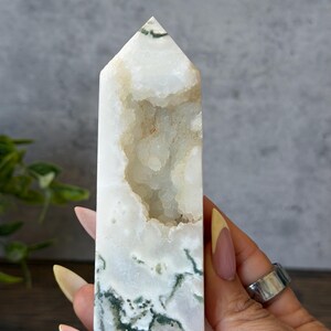 May include: A white and green crystal tower with a pointed top. The crystal has a mix of white, green, and tan colors. The crystal is held by a person.