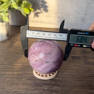 May include: A polished, spherical pink and white stone resting on a small circular stand. A black digital caliper is positioned around the sphere, displaying a measurement of 68.4 mm on its screen, indicating the diameter of the stone object.