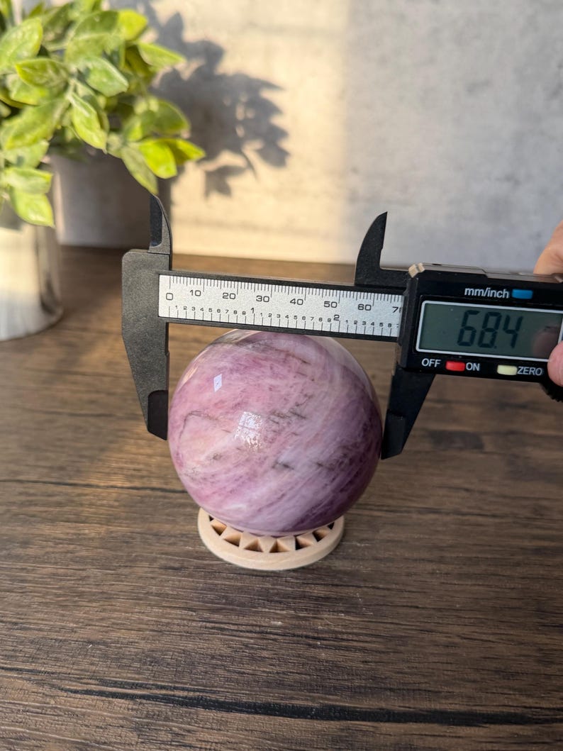 May include: A polished, spherical pink and white stone resting on a small circular stand. A black digital caliper is positioned around the sphere, displaying a measurement of 68.4 mm on its screen, indicating the diameter of the stone object.