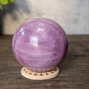 May include: A polished, spherical lavender-coloured stone orb with visible internal crystalline textures and swirling patterns. The sphere rests securely on a circular, light-coloured wooden stand featuring a series of small, triangular cutouts.