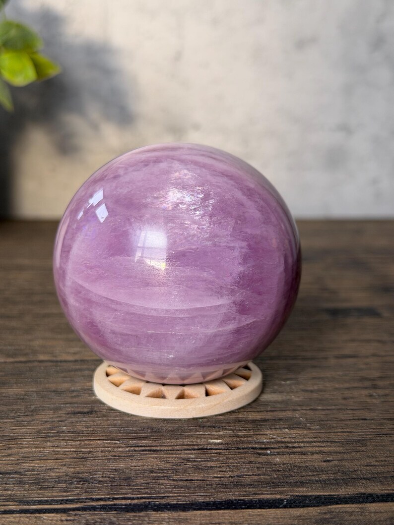 May include: A polished, spherical lavender-coloured stone orb with visible internal crystalline textures and swirling patterns. The sphere rests securely on a circular, light-coloured wooden stand featuring a series of small, triangular cutouts.