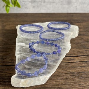 Puede incluir: Cinco pulseras de amatista p&uacute;rpura sobre una placa de carga de selenita blanca. Las pulseras est&aacute;n hechas de peque&ntilde;as cuentas redondas.