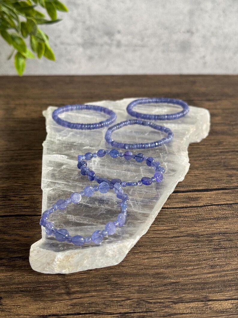 Puede incluir: Cinco pulseras de amatista p&uacute;rpura sobre una placa de carga de selenita blanca. Las pulseras est&aacute;n hechas de peque&ntilde;as cuentas redondas.