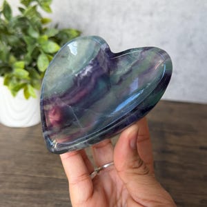 May include: A heart-shaped fluorite dish with a polished surface. The dish features a blend of green, purple, and blue hues. The heart-shaped dish is held by a hand, with a wooden surface in the background.