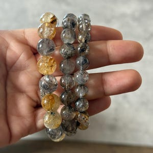 May include: Three beaded bracelets with clear, round beads. The beads have inclusions of gold, black, and gray. The bracelets are held in a hand, showcasing the natural variations in color and texture of the gemstones.