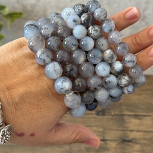 May include: A stack of gray and white gemstone bracelets with black flecks. The bracelets are made of round beads and are stacked on top of each other.
