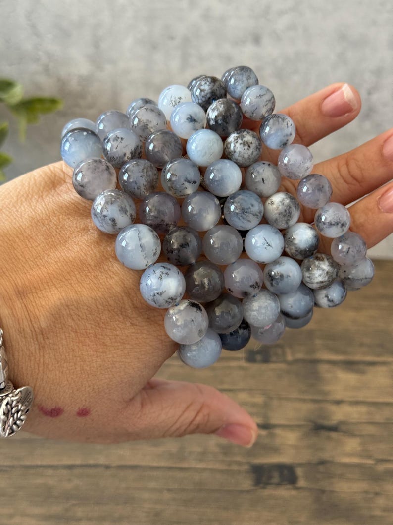 May include: A stack of gray and white gemstone bracelets with black flecks. The bracelets are made of round beads and are stacked on top of each other.
