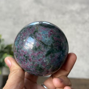 May include: A green and pink ruby zoisite sphere, a type of gemstone, is held in a hand. The sphere is smooth and polished, with a swirling pattern of green and pink.