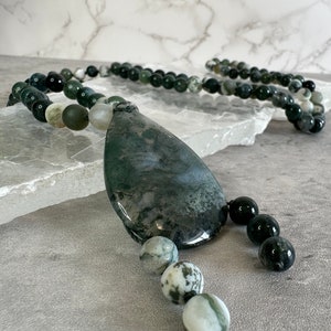 May include: A green and white agate gemstone necklace with a large, teardrop-shaped pendant. The necklace is made with round beads and has a black tassel.