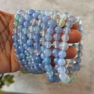 Puede incluir: Una pila de cuentas de piedra de &aacute;gata azul y blanca, cada cuenta es redonda y lisa. Las cuentas est&aacute;n enhebradas en un cord&oacute;n el&aacute;stico transparente.