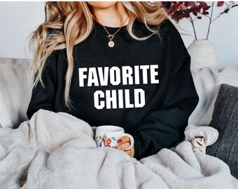 May include: A black sweatshirt with the text "FAVORITE CHILD" printed in white on the front. The sweatshirt is being worn by a person sitting on a couch with a blanket.