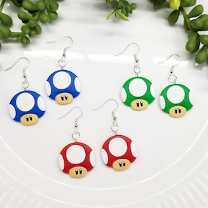 May include: A set of three pairs of mushroom-shaped earrings. Each pair features a different color: blue, green, and red. The earrings are made of polymer clay and have a white spot on top.
