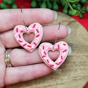 Op de afbeelding: Een paar roze hartvormige oorbellen met witte en rode sprinkles. De oorbellen zijn gemaakt van polymer clay en hebben een donut-achtig uiterlijk.