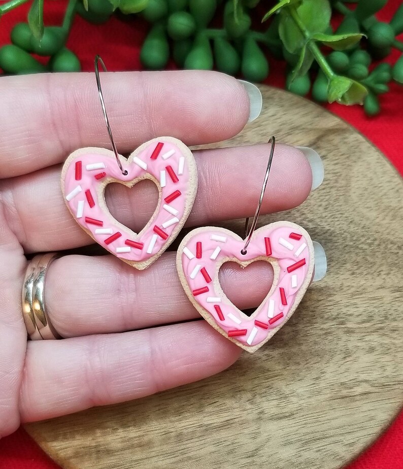 Op de afbeelding: Een paar roze hartvormige oorbellen met witte en rode sprinkles. De oorbellen zijn gemaakt van polymer clay en hebben een donut-achtig uiterlijk.