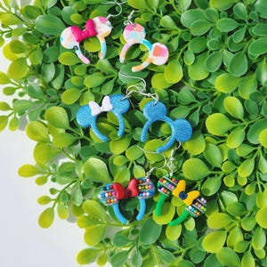 May include: A set of six pairs of colorful mouse ear earrings. The earrings are made of polymer clay and feature different designs, including polka dots, stripes, and glitter.