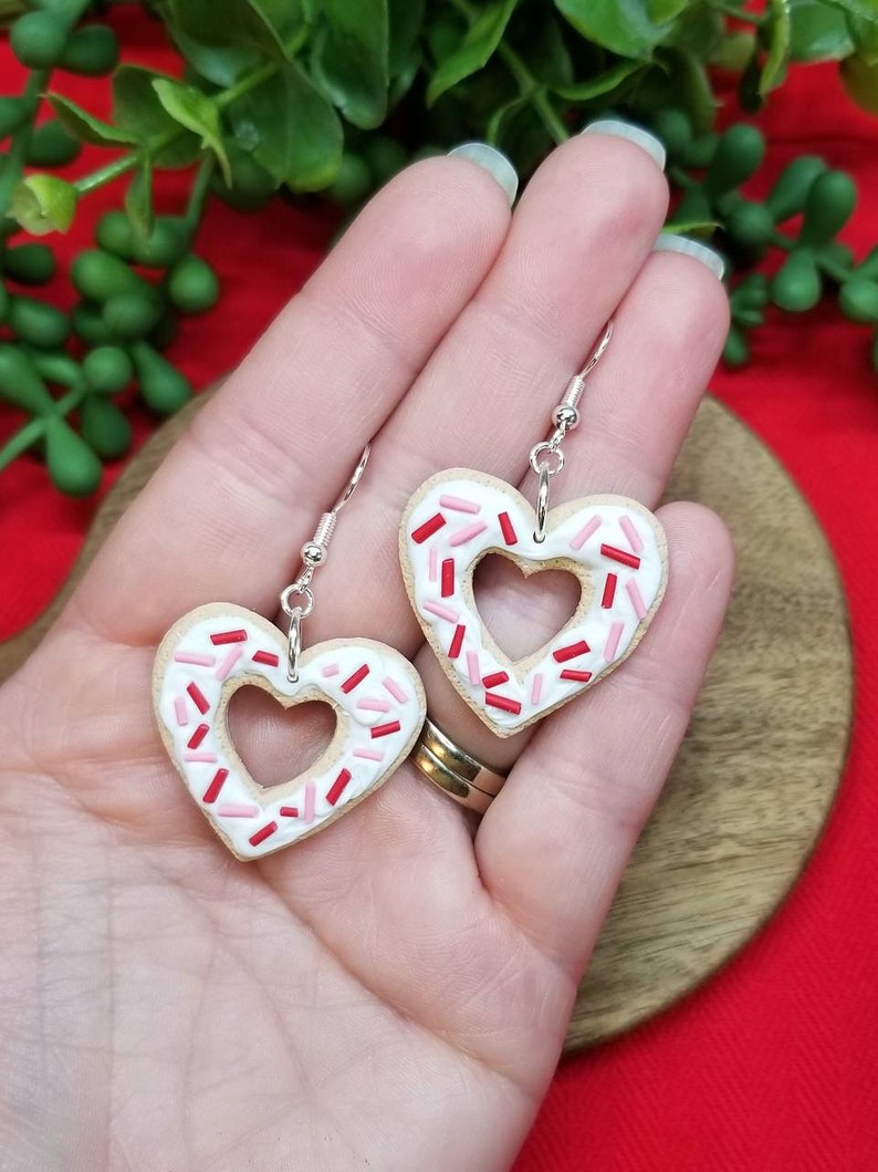 Op de afbeelding: Een paar hartvormige oorbellen gemaakt van witte polymer klei met roze en rode sprinkles. De oorbellen zijn aan een zilveren haak.