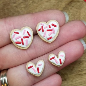 Op de afbeelding: Drie hartvormige suikerkoekjes met witte glazuur en rode en roze sprinkles. De koekjes zijn in een rij op een hand gerangschikt.
