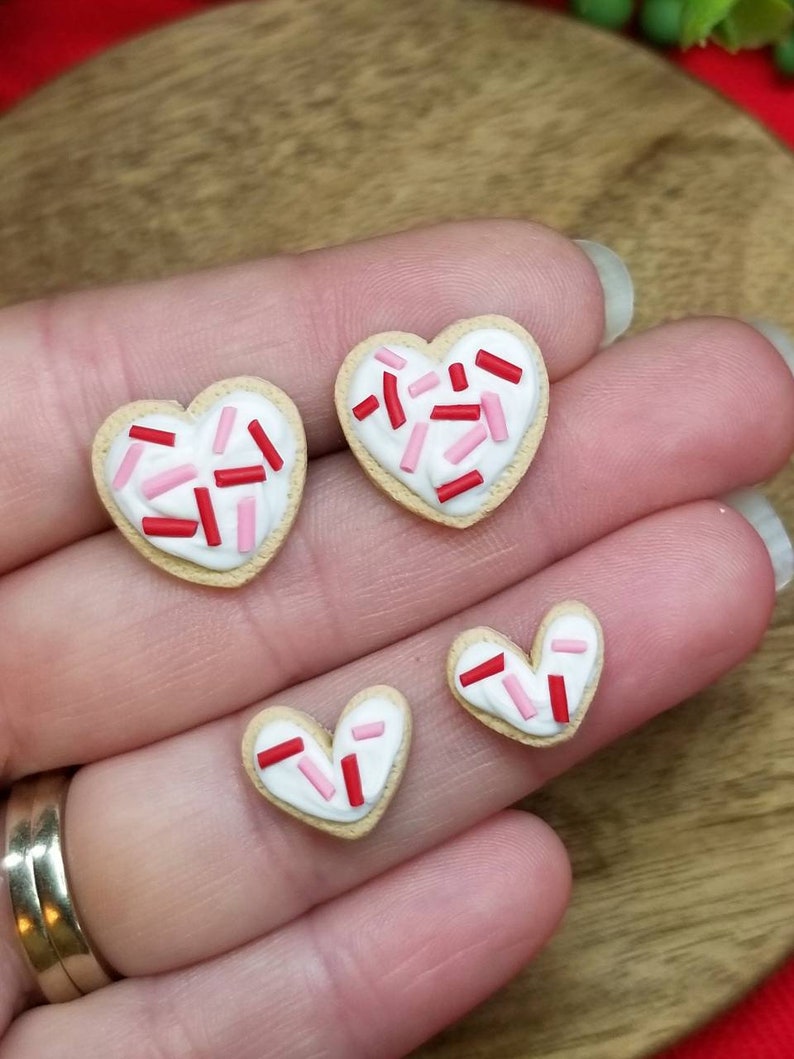 Op de afbeelding: Drie hartvormige suikerkoekjes met witte glazuur en rode en roze sprinkles. De koekjes zijn in een rij op een hand gerangschikt.