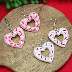 Op de afbeelding: Een paar hartvormige oorbellen met roze en witte frosting en rode en witte sprinkles. De oorbellen zijn gemaakt van polymer clay en hebben zilveren haakjes.