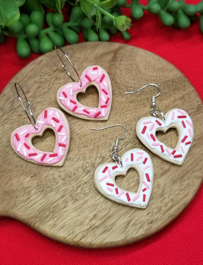 Op de afbeelding: Een paar hartvormige oorbellen met roze en witte frosting en rode en witte sprinkles. De oorbellen zijn gemaakt van polymer clay en hebben zilveren haakjes.