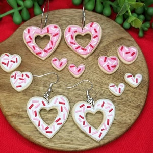 Op de afbeelding: Een set van zes hartvormige oorbellen gemaakt van polymer clay, versierd met roze en rode sprinkles. De oorbellen hebben de vorm van een hart met een gat in het midden. De oorbellen liggen op een houten oppervlak.