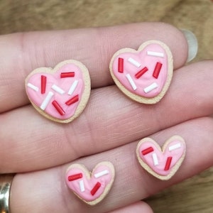 Op de afbeelding: Drie hartvormige koekjes met roze glazuur en rode en witte sprinkles. De koekjes zijn van verschillende grootte en zijn in een rij op een hand gerangschikt.