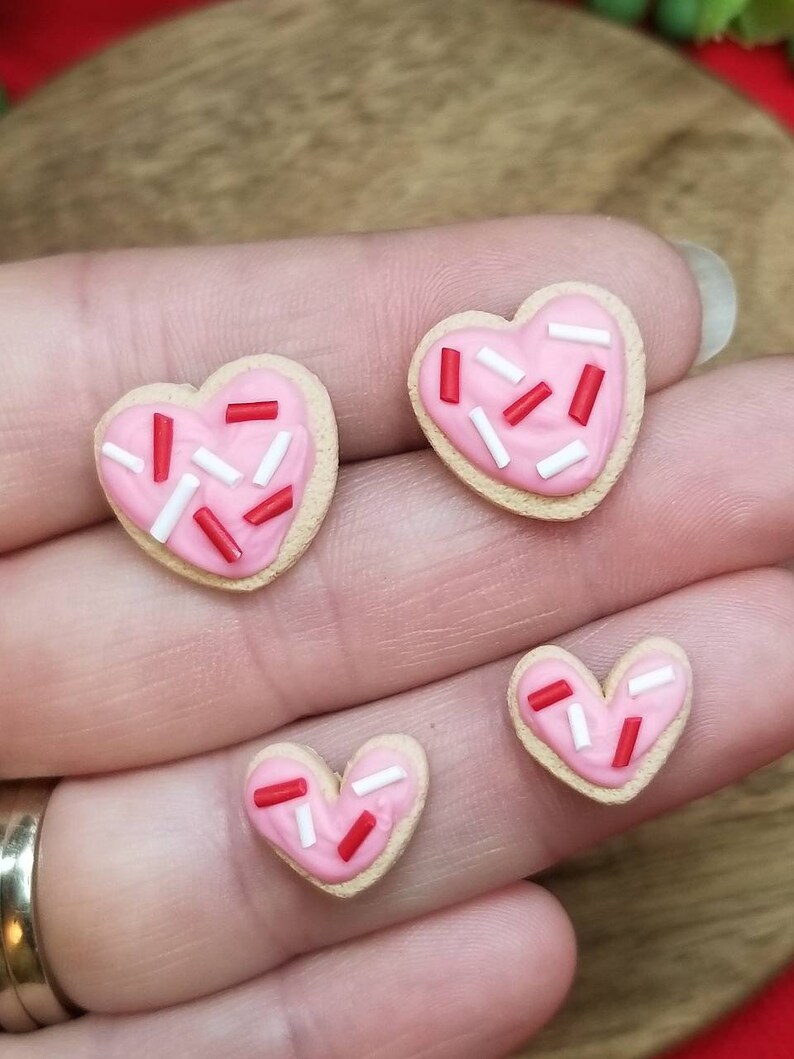 Op de afbeelding: Drie hartvormige koekjes met roze glazuur en rode en witte sprinkles. De koekjes zijn van verschillende grootte en zijn in een rij op een hand gerangschikt.